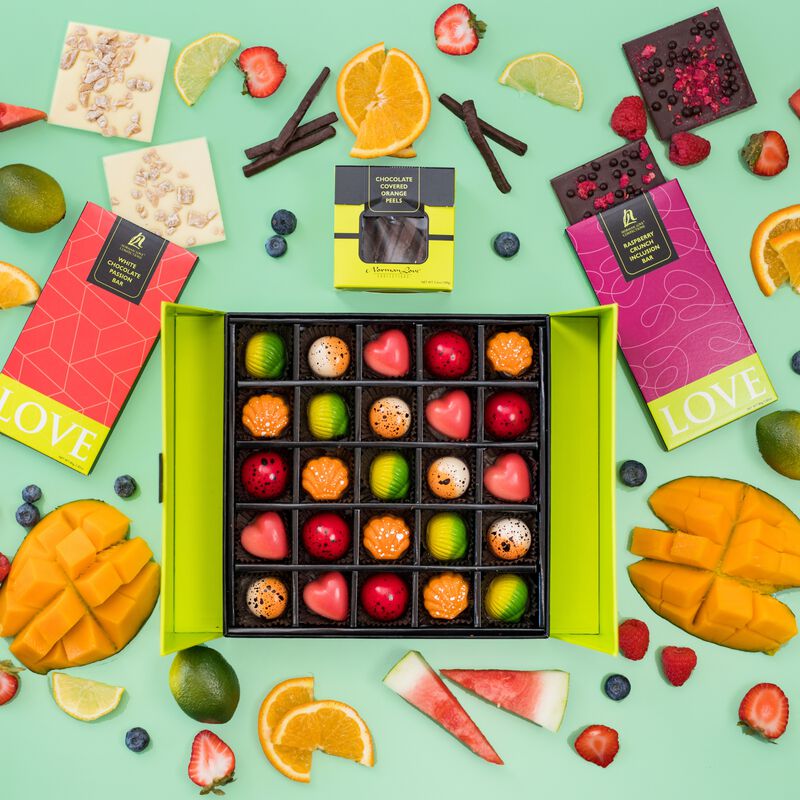 Overhead view of 25 piece gift box of fruit flavored truffles. Raspberry Crunch Chocolate Inclusion Bar, White Chocolate Passionfruit Inclusion Bar, and a package of Chocolate-covered Candied Orange Peels included in gift bundle. 